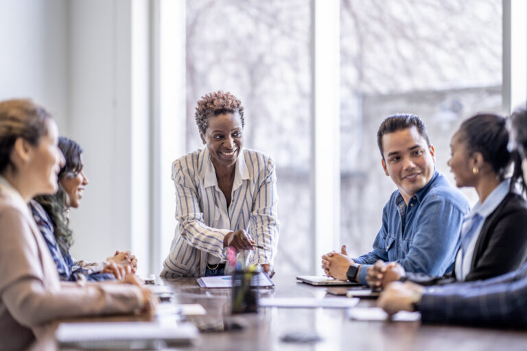 A small group of business professionals sit around a table as they meet to brainstorm some ideas for the future of the company. They are each dressed professionally and have papers scattered out in front of them as they work together. The focus is on a woman of African decent who is pitching an idea to the group.